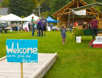 A photo of a farmers market, with grass in the foreground, tents, a few people, and a wooden pavilion in the background. A hand painted sign in the front next to a wooden bridge reads "Welcome! We are so glad you are here." 