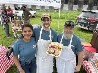 Three CAE staff members, outdoors holding up a container with a local burger, slaw, and potatoes. 